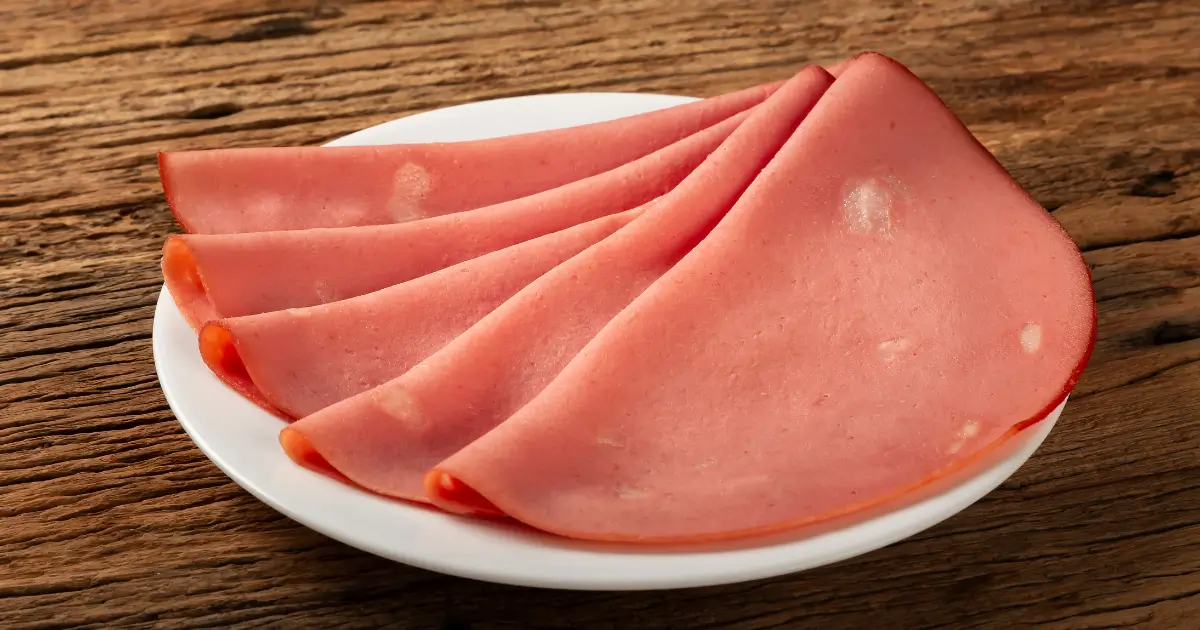 Fatias finas de mortadela dispostas sobre um prato branco, que está colocado em uma superfície de madeira rústica. As fatias têm uma cor rosa característica, com algumas pequenas manchas brancas, típicas de gordura, visíveis na mortadela.
