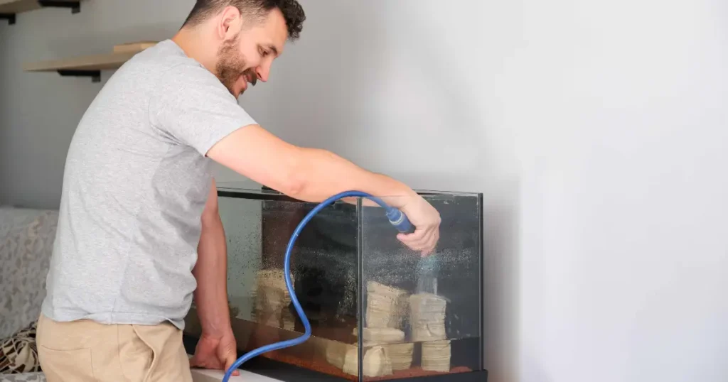 A imagem mostra um homem limpando um aquário. Ele está utilizando um sifão ou mangueira de sucção para remover a água do tanque, possivelmente para uma manutenção ou troca de água. O homem sorri enquanto realiza o procedimento, segurando firmemente a mangueira dentro do aquário, que possui um substrato no fundo e algumas pedras ou decorações. A cena sugere um ambiente doméstico, com o aquário posicionado contra uma parede clara.