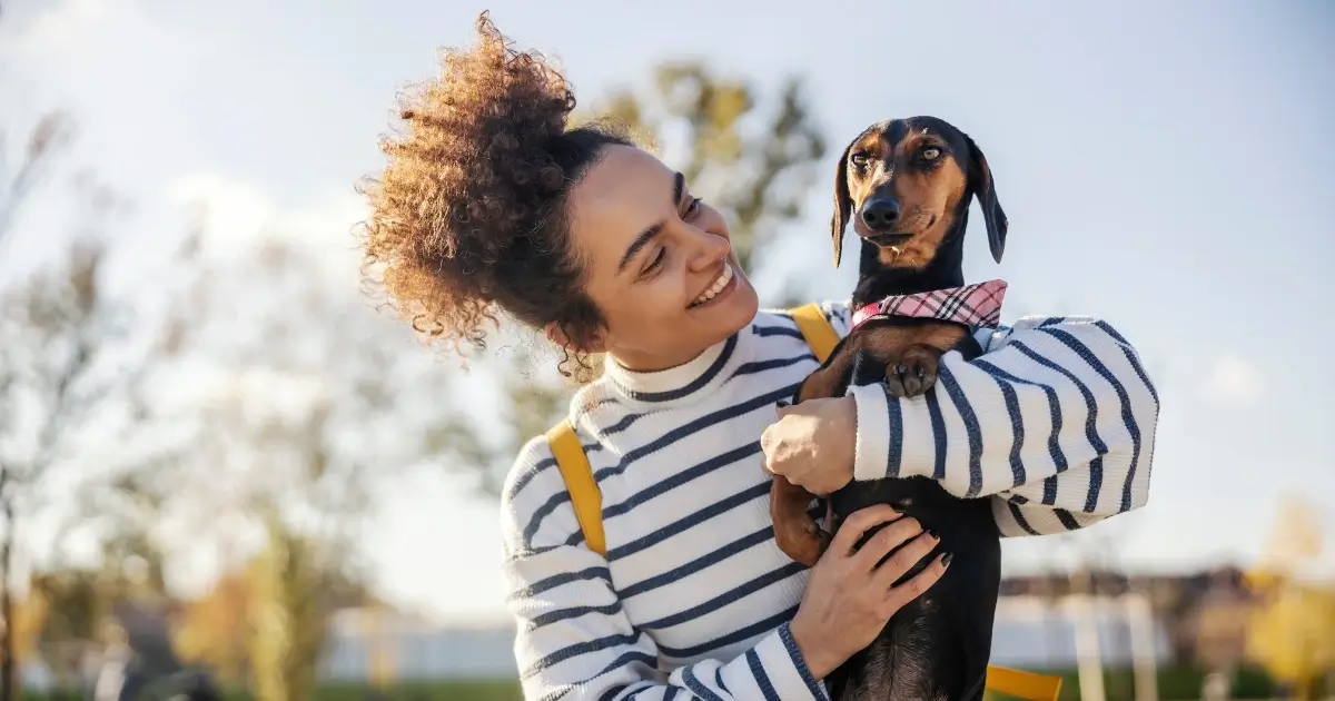 A imagem mostra uma mulher sorridente segurando um cachorro da raça Dachshund (salsicha) nos braços, ao ar livre. Ela veste um suéter listrado branco e azul, com alças de mochila amarelas visíveis. O cachorro usa um lenço rosa xadrez no pescoço e tem uma expressão calma e observadora. O fundo desfocado revela um ambiente ensolarado, com árvores e céu azul, sugerindo um dia agradável ao ar livre.