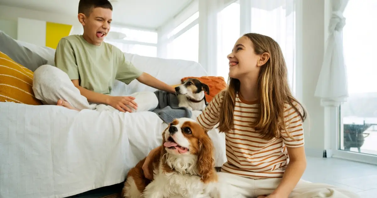 A imagem mostra duas crianças, um menino e uma menina, interagindo com dois cachorros dentro de um ambiente doméstico bem iluminado. O menino, sentado no sofá, veste uma camiseta verde clara e segura um dos cães, que está parcialmente coberto por um cobertor. A menina, sentada no chão, usa uma blusa listrada e sorri enquanto acaricia o outro cachorro, um Cavalier King Charles Spaniel, que parece feliz e relaxado. O cenário transmite um clima acolhedor e harmonioso, sugerindo um ambiente adequado para pets em apartamentos.
