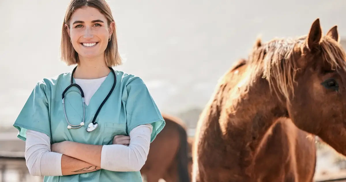 A imagem mostra uma veterinária sorridente vestindo jaleco verde e um estetoscópio ao redor do pescoço, de braços cruzados, transmitindo confiança e profissionalismo. Ao fundo, um cavalo marrom está parcialmente visível, indicando que ela trabalha com animais de grande porte, possivelmente em uma fazenda ou clínica especializada em equinos. O cenário é ao ar livre, com iluminação natural, reforçando a conexão entre a profissional e os animais que cuida.